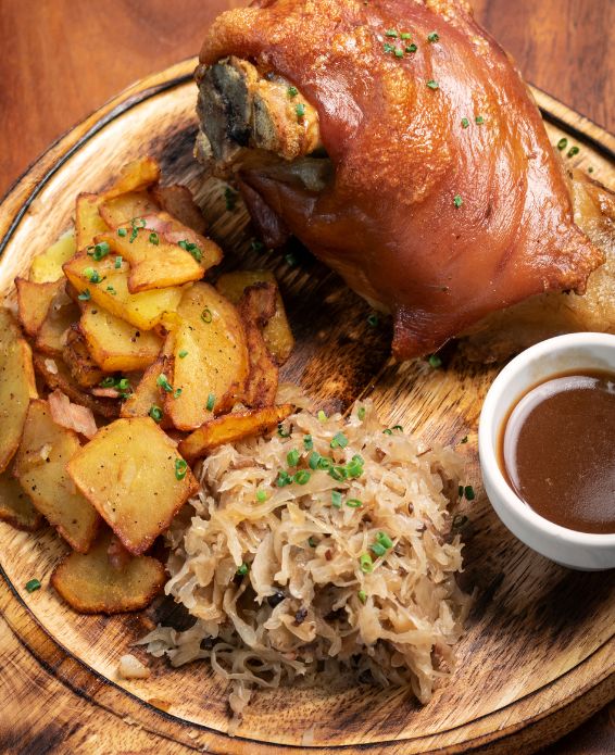 Rustikales Gericht mit knuspriger Schweinshaxe auf einem Holzbrett, serviert mit Bratkartoffeln mit Speck, Sauerkraut und dunkler Bratensoße in einem kleinen Schälchen – garniert mit frischem Schnittlauch.